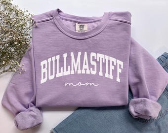 Może przedstawiać: Liliowa bluza z napisem "BULLMASTIFF" w białych, blokowych literach i słowem "mom" kursywą. Bluza ma okrągły dekolt i długie rękawy z podwiniętymi mankietami. Bluza jest złożona na parze niebieskich dżinsów.