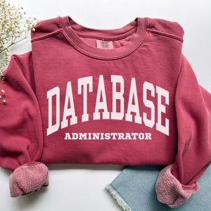 Könnte beinhalten: Ein weinrotes Sweatshirt mit dem weißen Aufdruck "DATABASE ADMINISTRATOR". Das Sweatshirt hat einen Rundhalsausschnitt und gerippte Bündchen. Die Ärmel sind hochgekrempelt. Das Kleidungsstück ist tiefrot.