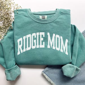 May include: A teal-colored sweatshirt with the text "RIDGIE MOM" in white block letters. The sweatshirt has a crew neck and long sleeves with ribbed cuffs. The brand tag "COMFORT COLORS" is visible on the upper back.