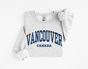 May include: A light gray crewneck sweatshirt featuring the text VANCOUVER in a large, dark blue arched font, with the word CANADA printed in a smaller, straight dark blue font directly underneath the city name on the front of the garment.