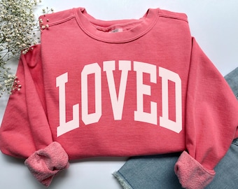 Könnte beinhalten: Ein korallenfarbenes Sweatshirt mit dem Wort "LOVED" in großen, weißen Blockbuchstaben auf der Vorderseite. Das Sweatshirt hat einen Rundhalsausschnitt und gerippte Bündchen. Die Ärmel sind umgeschlagen und der Stoff wirkt weich und bequem.