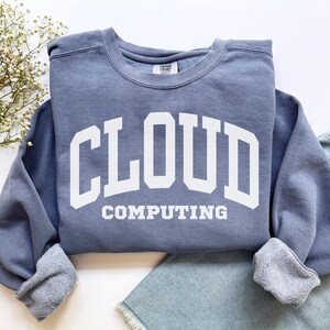 Könnte beinhalten: Ein blaues Sweatshirt mit den Worten "CLOUD COMPUTING" in großen weißen Blockbuchstaben. Das Sweatshirt hat einen Rundhalsausschnitt und gerippte Bündchen. Das Kleidungsstück ist auf einer blauen Jeans gefaltet.