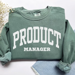 May include: A sage green sweatshirt with the words "PRODUCT MANAGER" in large, white, block letters. Below, in smaller white letters, is the word "MANAGER". The sweatshirt has a crew neck and ribbed cuffs.