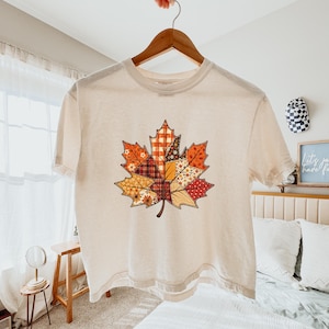 Puede incluir: Una camiseta de manga corta de color crema con un gráfico central grande de una hoja de arce. El diseño de la hoja está compuesto por varios patrones de estilo patchwork en tonos naranja, rojo, amarillo y marrón, incluyendo cuadros y flores.