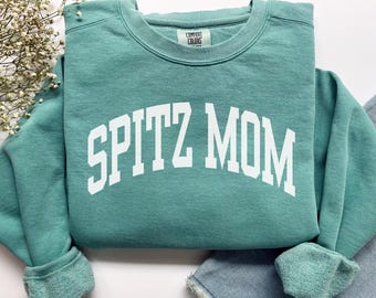 Może przedstawiać: Teal bluza Comfort Colors z napisem "SPITZ MOM" w białych literach blokowych. Bluza ma okrągły dekolt i długie rękawy ze ściągaczami. Odzież jest złożona na parze niebieskich dżinsów.