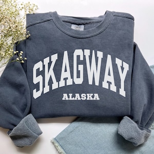 Puede incluir: Una sudadera de cuello redondo gris carbón con el texto blanco SKAGWAY en una fuente universitaria arqueada y la palabra ALASKA impresa debajo en una fuente más pequeña y recta. La prenda tiene mangas largas y un interior de forro polar.