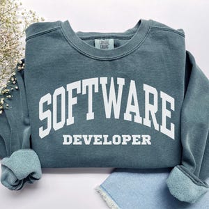 May include: A teal sweatshirt with the words "SOFTWARE DEVELOPER" in white block letters. The sweatshirt has a crew neck and ribbed cuffs and hem. The Comfort Colors brand tag is visible.