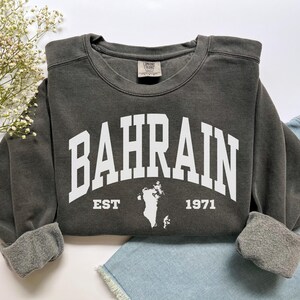 May include: Dark gray sweatshirt with the word "BAHRAIN" in large white block letters. Below the word is "EST 1971" and a white outline of the country. The sweatshirt has a crew neck and long sleeves with cuffed wrists.