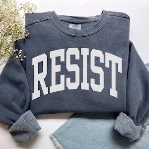 May include: A dark blue sweatshirt with the word "RESIST" printed in large, white, block letters across the front. The sweatshirt has a crew neck and cuffed sleeves. The garment is folded on a white surface with a sprig of white flowers and a piece of denim.