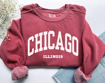 May include: A maroon sweatshirt with the word "CHICAGO" in large white block letters arched across the chest, with "ILLINOIS" below. The sleeves are cuffed, and the sweatshirt is on a white surface with a sprig of white flowers and a pair of blue jeans.