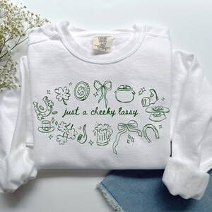 May include: A white crewneck sweatshirt featuring a dark green line art graphic with the text 'just a cheeky lassy'. The design includes illustrations of shamrocks, horseshoes, a pot of gold, a beer mug, a pipe, bows, and a leprechaun hat.
