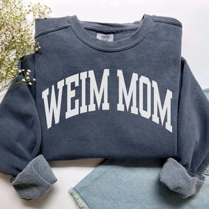 May include: A slate blue sweatshirt with the text "WEIM MOM" in white block letters. The sweatshirt has a crew neck and cuffed sleeves. The garment is displayed on a white surface with a sprig of white flowers and denim shorts.
