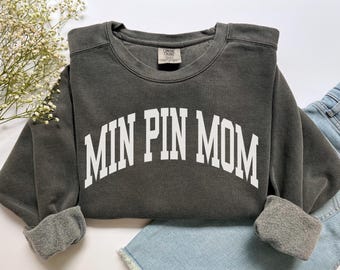 May include: Dark grey sweatshirt featuring the white text "MIN PIN MOM" in an arched design. The crew neck sweatshirt has cuffed sleeves and is folded on blue denim shorts. The garment is made of a soft material.