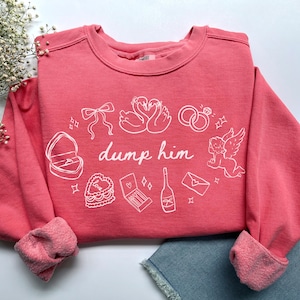 Puede incluir: Una sudadera de color coral con ilustraciones gráficas blancas y el texto "dump him". El diseño incluye una caja de anillos, cisnes, anillos de boda, un pastel, un cupido y otros símbolos románticos. Las mangas están dobladas.