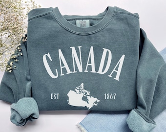 Pode incluir: Uma camisola de gola redonda azul ardósia com o texto branco CANADA numa fonte arqueada sobre um mapa de silhueta branca do Canadá, com o texto EST 1867 posicionado abaixo do mapa. A etiqueta interior do pescoço diz Comfort Colors.