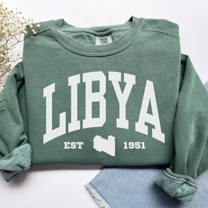 May include: A sage green sweatshirt with the word "LIBYA" in large white block letters. Below the word "LIBYA" is the text "EST 1951" and a white outline of the country of Libya. The sweatshirt has a crew neck and long sleeves.