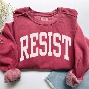 May include: A maroon sweatshirt with the word "RESIST" printed in large, white, block letters. The sweatshirt has a crew neck and ribbed cuffs. The sleeves are rolled up, and the garment is folded on a light background.