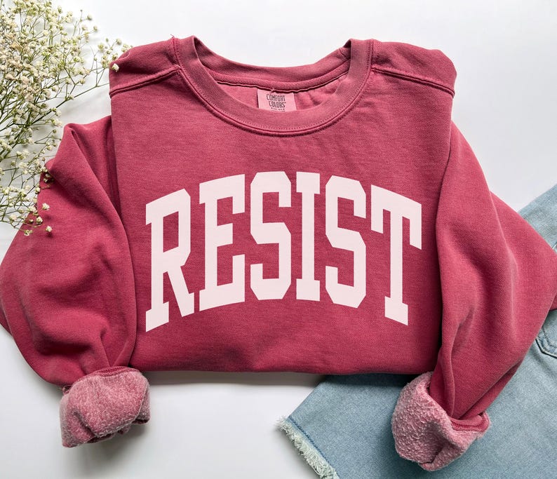 May include: A maroon sweatshirt with the word "RESIST" printed in large, white, block letters. The sweatshirt has a crew neck and ribbed cuffs. The sleeves are rolled up, and the garment is folded on a light background.