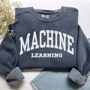 Puede incluir: Una sudadera azul pizarra con las palabras "MACHINE LEARNING" impresas en letras blancas. La sudadera tiene cuello redondo y puños. Se muestra sobre una superficie blanca con una ramita de flores blancas y un par de shorts vaqueros azules.