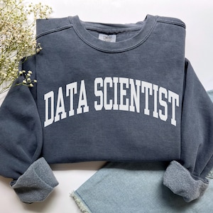 Könnte beinhalten: Ein dunkelgraues Sweatshirt mit dem weißen Schriftzug "DATA SCIENTIST" in bogenförmigen Buchstaben. Das Sweatshirt hat einen Rundhalsausschnitt und gerollte Ärmel. Das Kleidungsstück wird auf einer weißen Oberfläche mit einem Zweig weißer Blumen und Jeansshorts präsentiert.