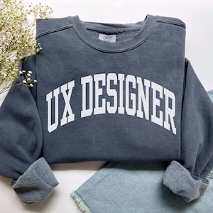 May include: A dark grey sweatshirt featuring the white text 'UX DESIGNER' in a bold, arched font. The crew neck sweatshirt has cuffed sleeves and is displayed on a white surface, alongside a sprig of white flowers and denim shorts.