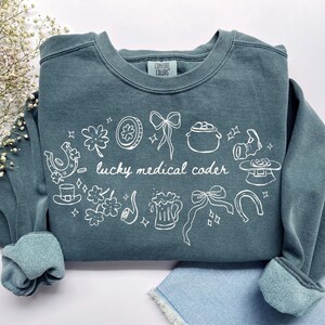 May include: A slate blue crewneck sweatshirt featuring a white line art graphic with the text "lucky medical coder" surrounded by icons including shamrocks, a pot of gold, horseshoes, ribbons, a beer mug, and a top hat, with a Comfort Colors label.