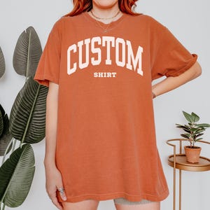 以下が含まれることがあります： 胸に白いアーチ状の文字で「CUSTOM」、下に「SHIRT」と書かれた、錆色のTシャツ。半袖でゆったりとしたフィット感です。
