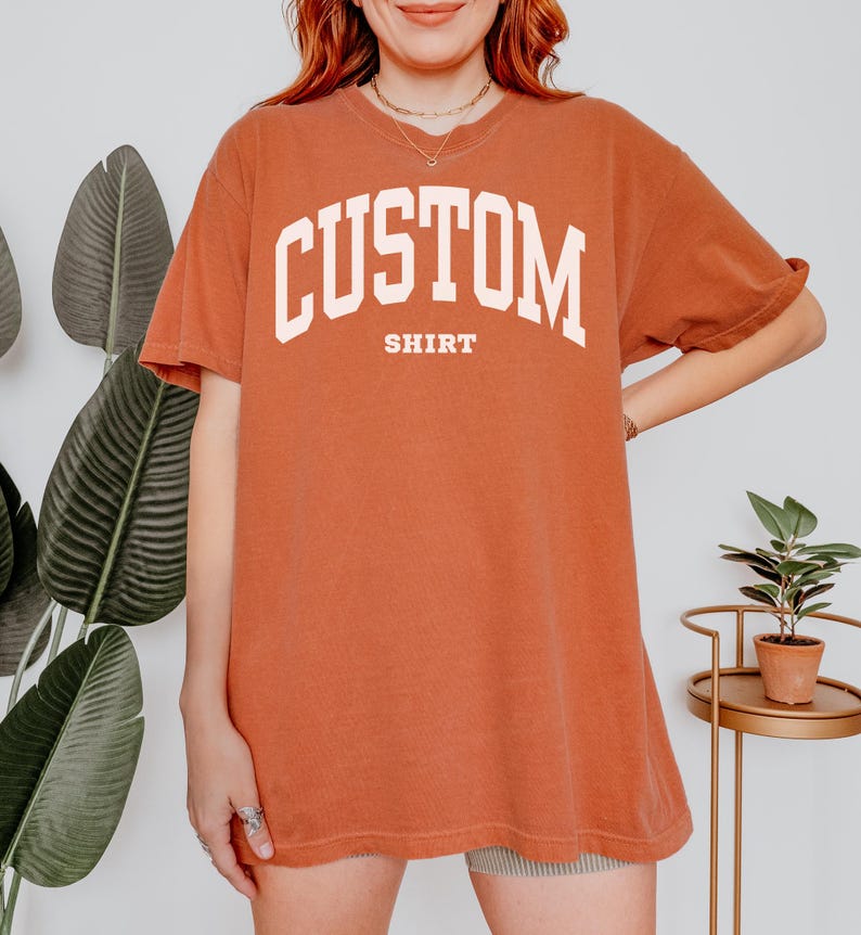 以下が含まれることがあります： 胸に白いアーチ状の文字で「CUSTOM」、下に「SHIRT」と書かれた、錆色のTシャツ。半袖でゆったりとしたフィット感です。