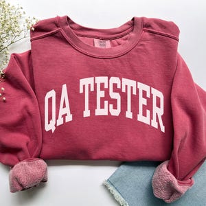 Può includere: Una felpa color bordeaux con la scritta bianca "QA TESTER". La felpa ha girocollo, maniche lunghe e un aspetto morbido e strutturato. I polsini e il colletto hanno un aspetto sbiadito e vintage.
