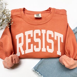 May include: A burnt orange sweatshirt with the word "RESIST" in large, white, block letters across the front. The sweatshirt is folded over a pair of light blue denim shorts. The sleeves are cuffed, revealing a lighter orange interior.