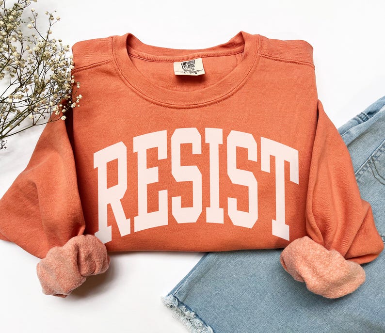 May include: A burnt orange sweatshirt with the word "RESIST" in large, white, block letters across the front. The sweatshirt is folded over a pair of light blue denim shorts. The sleeves are cuffed, revealing a lighter orange interior.