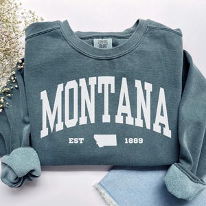 Könnte beinhalten: Ein blaugrüner Pullover mit dem Wort "MONTANA" in weißen Blockbuchstaben, mit "EST" und "1889" auf beiden Seiten einer weißen Umrandung des Bundesstaates Montana.