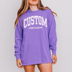 以下が含まれることがあります： 胸に白いブロック体で「CUSTOM」の文字がアーチ状にプリントされた長袖紫シャツ。「LONG SLEEVE」の文字が下に白でプリントされています。ゆったりとしたフィット感のシャツです。