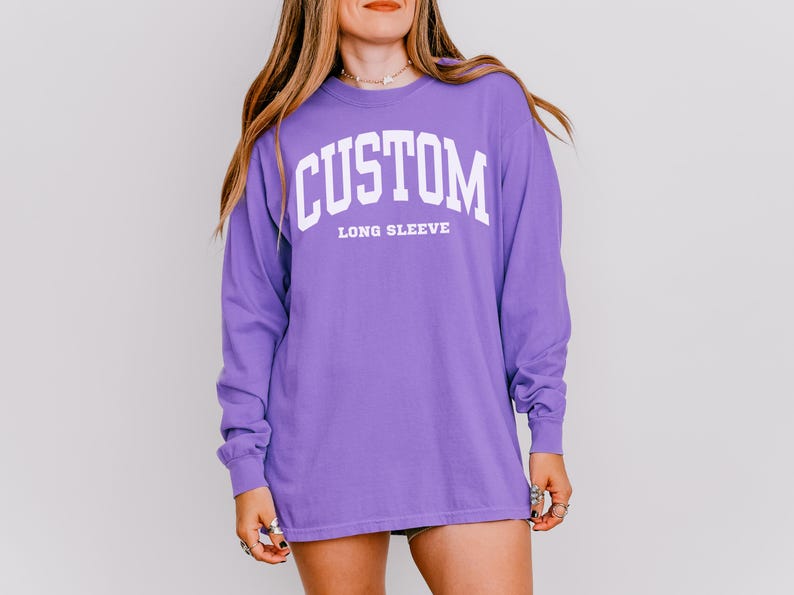 以下が含まれることがあります： 胸に白いブロック体で「CUSTOM」の文字がアーチ状にプリントされた長袖紫シャツ。「LONG SLEEVE」の文字が下に白でプリントされています。ゆったりとしたフィット感のシャツです。