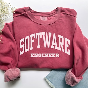 Puede incluir: Una sudadera granate con las palabras "SOFTWARE ENGINEER" en letras blancas. La sudadera tiene cuello redondo y mangas largas con puños enrollados. La prenda está hecha de un material suave.