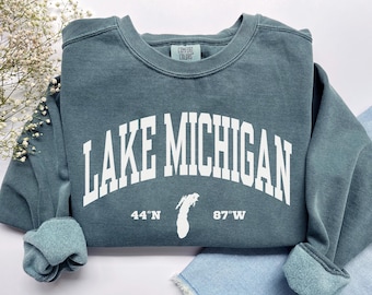 May include: A slate blue crewneck sweatshirt featuring the white text "LAKE MICHIGAN" in an arched collegiate font. Below the text is a white silhouette of Lake Michigan flanked by the coordinates "44°N" and "87°W". The inner label reads "COMFORT COLORS".
