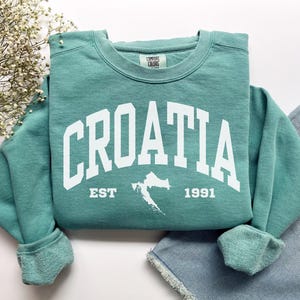 Puede incluir: Sudadera de cuello redondo color turquesa con la palabra "CROATIA" en letras blancas. Debajo, un mapa de Croacia y el texto "EST 1991". La sudadera está doblada, con unos vaqueros visibles en el fondo.