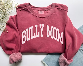 Może przedstawiać: Ciemnoróżowa bluza z okrągłym dekoltem z napisem BULLY MOM wykonanym białymi wielkimi literami, ułożonymi w łuk na klatce piersiowej. Odzież posiada długie rękawy ze ściągaczami oraz widoczną metkę marki wewnątrz dekoltu.