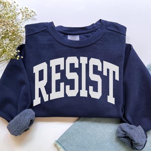 May include: A navy blue sweatshirt with the word "RESIST" in large, white, block letters across the front. The sweatshirt has a crew neck and cuffed sleeves. The garment is folded, with a portion of denim jeans visible.