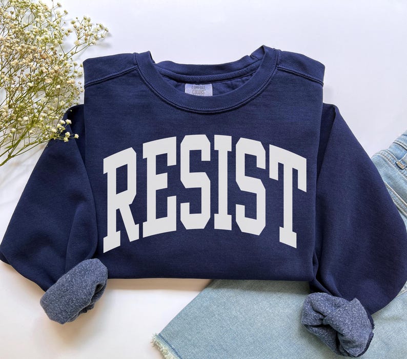 May include: A navy blue sweatshirt with the word "RESIST" in large, white, block letters across the front. The sweatshirt has a crew neck and cuffed sleeves. The garment is folded, with a portion of denim jeans visible.