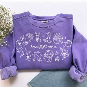 May include: A purple sweatshirt with white illustrations of Easter-themed drawings, including a bunny, duck, and cupcakes. The text "hoppy flight nurse" is printed in white. The sweatshirt has rolled-up sleeves.