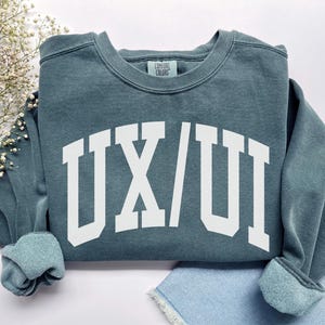 May include: A teal crewneck sweatshirt with the white text "UX/UI" in a collegiate font. The sweatshirt has ribbed cuffs and a ribbed hem. The brand "Comfort Colors" is printed on the back of the neck.