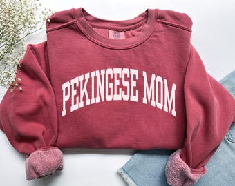 Może przedstawiać: Bordowa bluza z okrągłym dekoltem z białym napisem "PEKINGESE MOM". Bluza ma luźny krój z prążkowanymi mankietami i miękkim, wyblakłym wyglądem. Rękawy są podwinięte, odsłaniając jaśniejszą różową podszewkę.