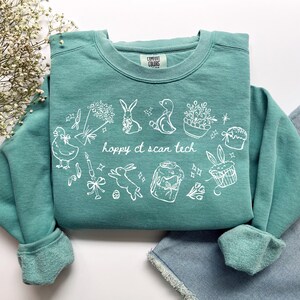 Peut inclure: Sweat-shirt de couleur sarcelle avec des illustrations blanches sur le thème de Pâques, notamment des lapins, des poussins et des carottes. Le texte "hoppy ct scan tech" est imprimé en blanc. Le sweat-shirt est en matière douce.