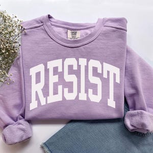 May include: A light purple sweatshirt with the word "RESIST" in large white block letters. The sweatshirt is folded, with the sleeves cuffed. A pair of blue jeans is visible in the background.