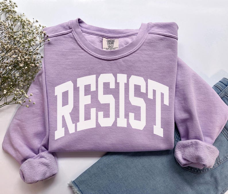 May include: A light purple sweatshirt with the word "RESIST" in large white block letters. The sweatshirt is folded, with the sleeves cuffed. A pair of blue jeans is visible in the background.