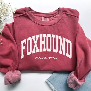 May include: A maroon sweatshirt with the text "FOXHOUND mom" in white block letters. The sweatshirt has a crew neck and cuffed sleeves. The cuffs and the inside of the collar are a lighter shade of maroon.
