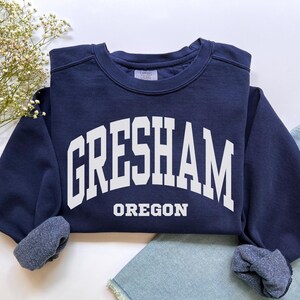 Puede incluir: Una sudadera azul marino con la palabra "Gresham" en letras blancas grandes arqueadas sobre el pecho, con "Oregon" debajo. La sudadera tiene cuello redondo y puños. La sudadera está doblada sobre una superficie blanca.