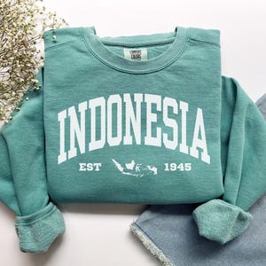 May include: Teal crew neck sweatshirt featuring the word "INDONESIA" in white block letters. Below the word is "EST 1945" and a white graphic of the Indonesian archipelago. The sweatshirt is folded, with the sleeves and bottom hem visible.