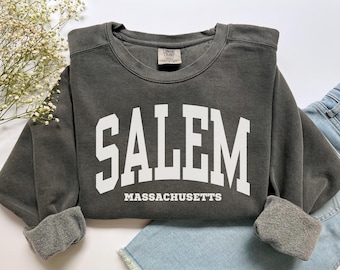Puede incluir: Sudadera gris oscuro con la palabra "SALEM" en letras blancas grandes y "MASSACHUSETTS" debajo. La sudadera tiene cuello redondo y mangas con puños. La prenda se muestra sobre una superficie de mezclilla azul claro.